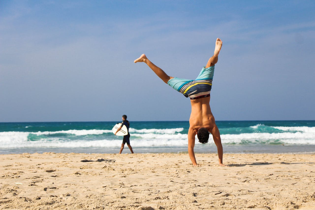 hero Man doing a handstand on a sunny beach with surfer in background, evoking a sense of freedom and joy.
